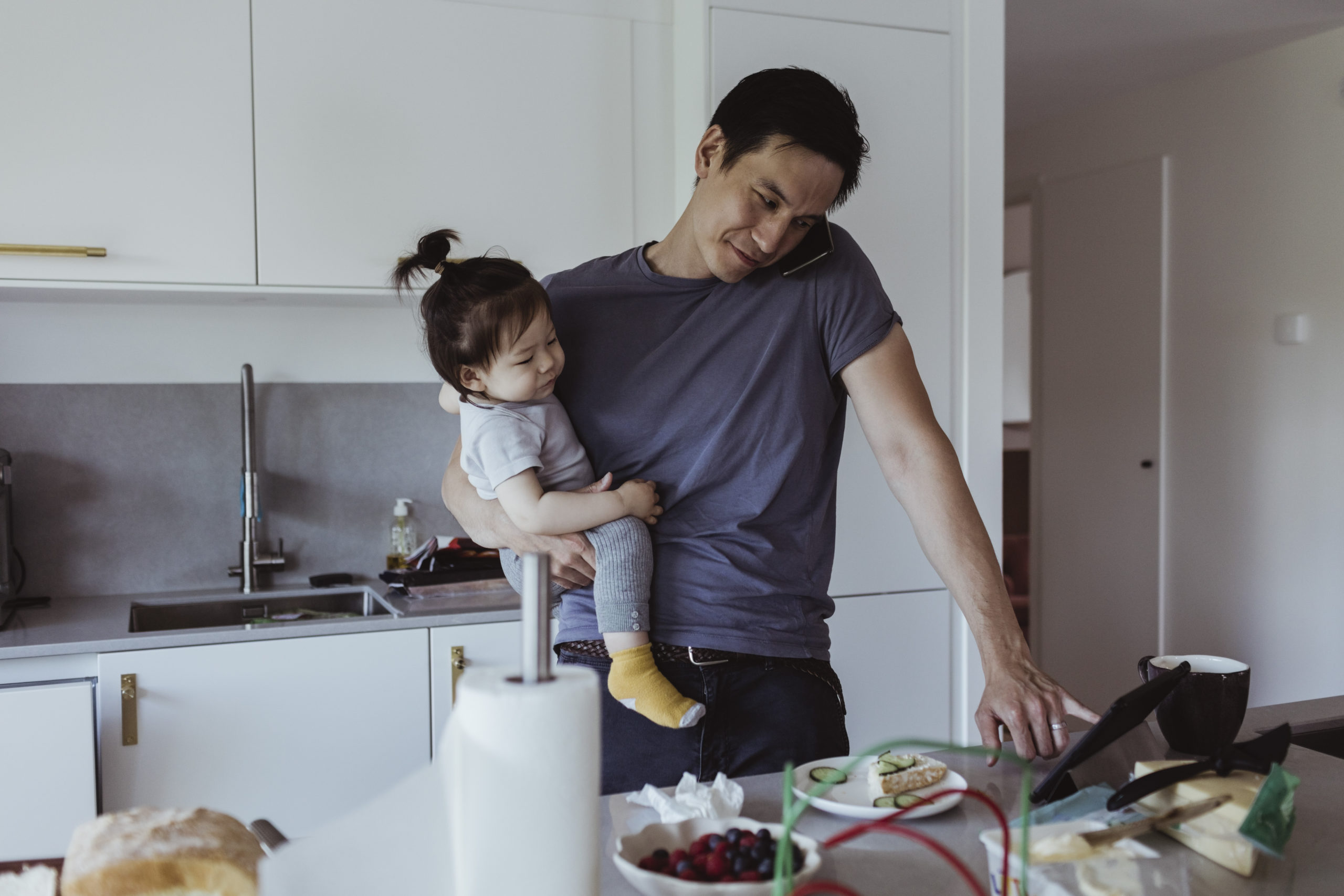 Father talking on smart phone while using digital tablet while carrying baby son in kitchen