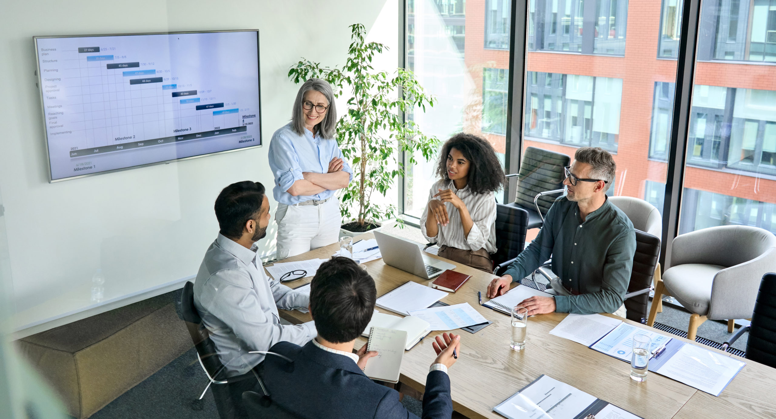 Senior female ceo and multicultural business people discussing pay transparency laws at boardroom table. Diverse corporate team working together in modern meeting room office. 