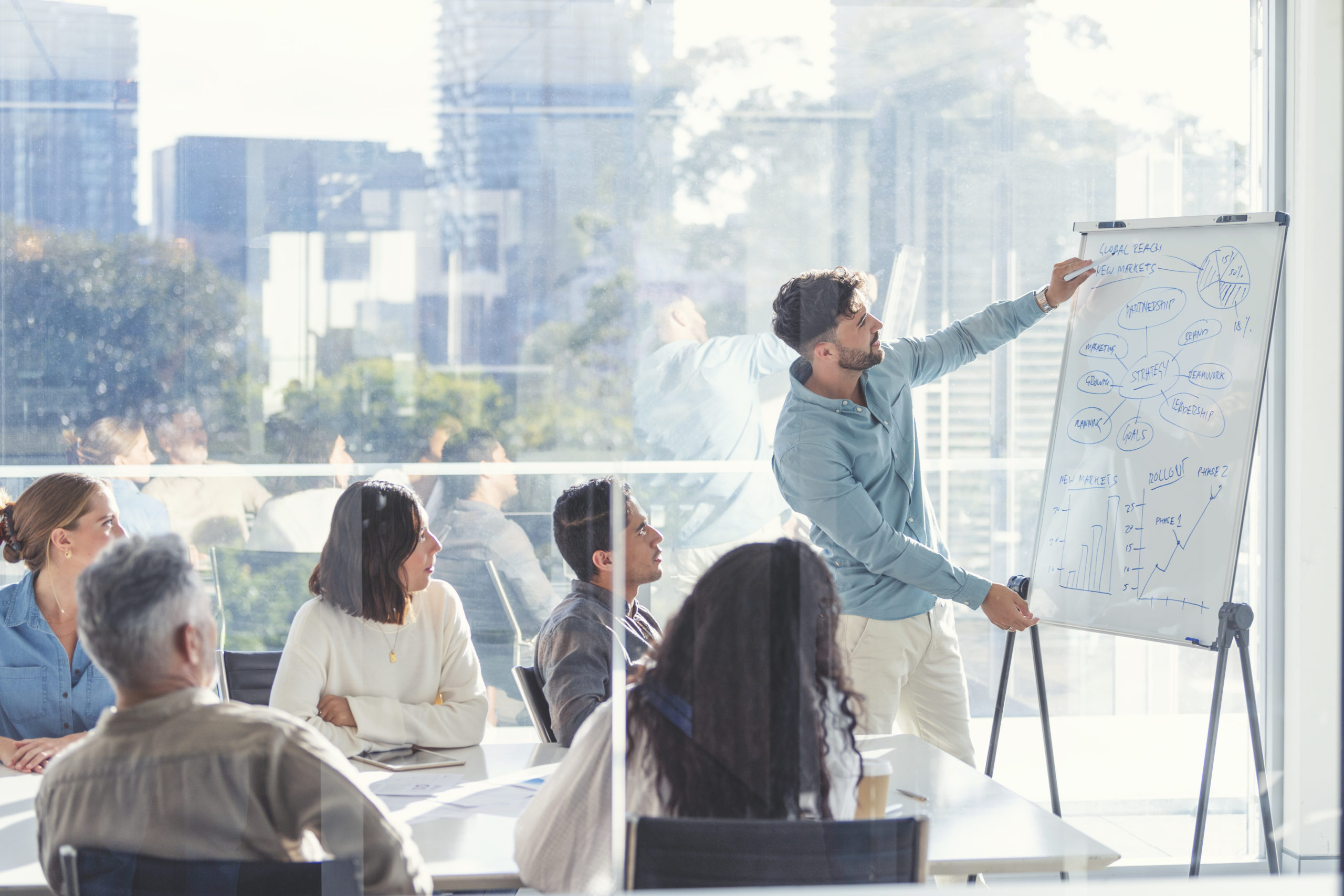 Business people watching a presentation on the whiteboard. An outsourced HR consultant is writing on the whiteboard with charts and graphs. They are sitting in a board room, there are laptop computers and technology on the table. All are casually dressed. There is a window behind him with city views.