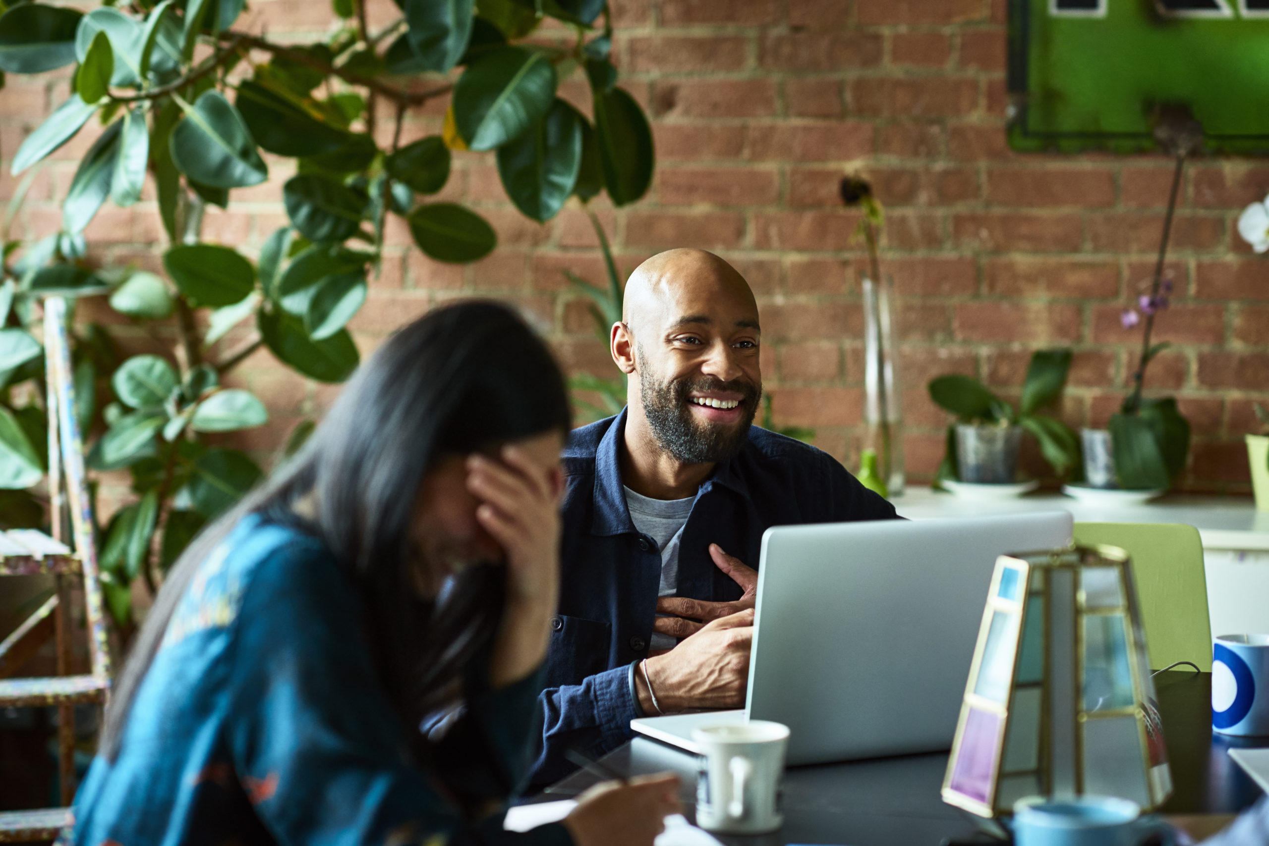 Mid adult man with laptop, smiling and looking away with woman in foreground, head in hands, laughing, carefree creative professionals discussing HR trends for 2023.