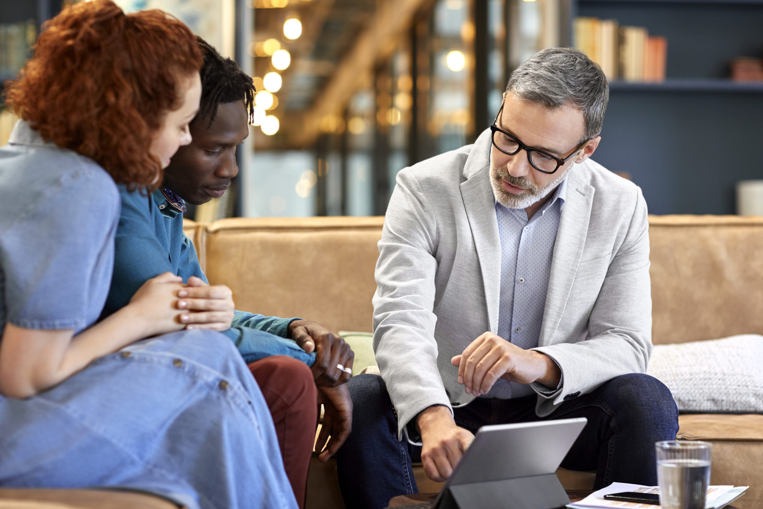 Mature financial advisor planning with young multi-ethnic couple at office. Businessman is discussing over digital tablet with clients at workplace. They are sitting in meeting.