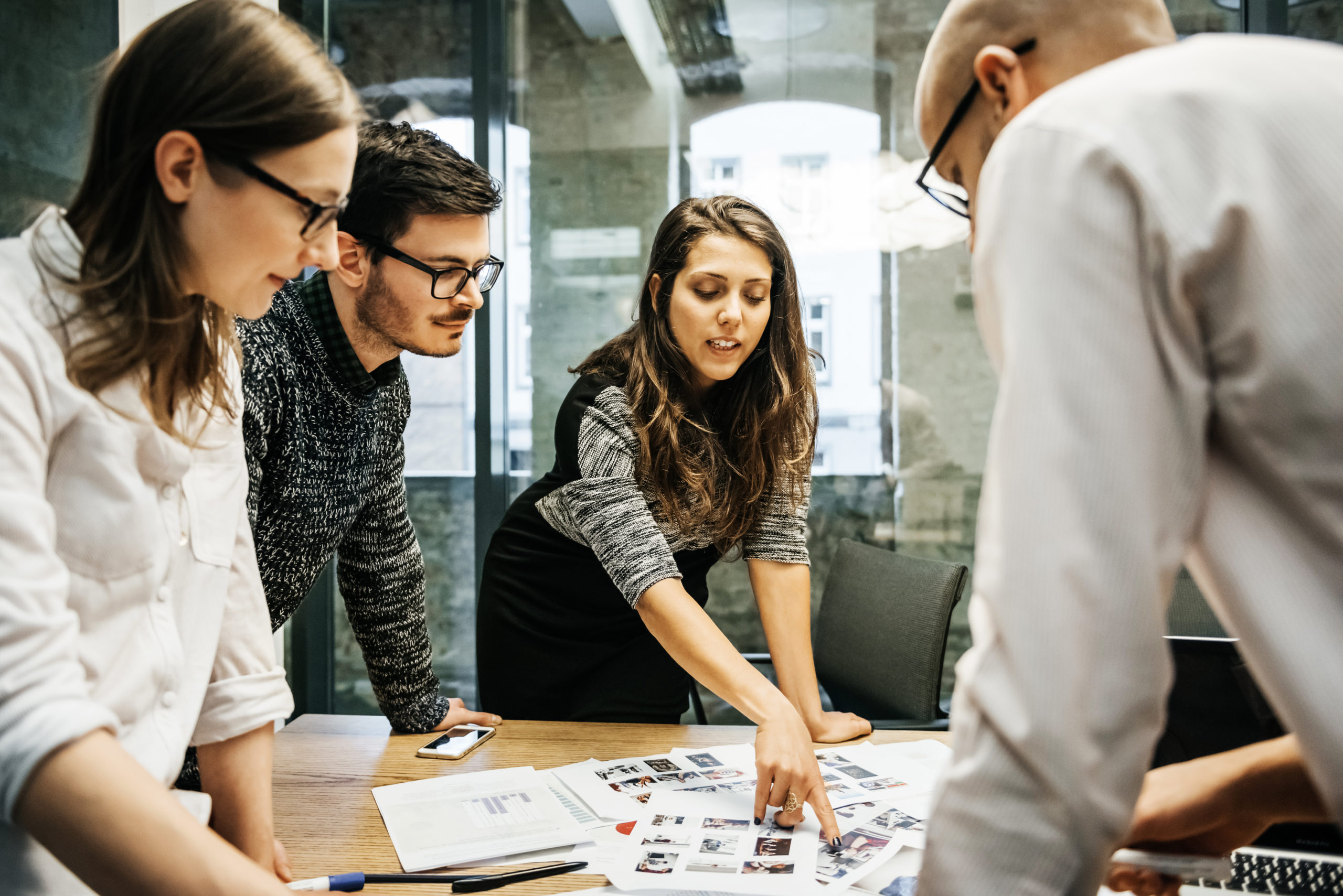 A team of young people in a modern office is discussing their project. The group consists of two men and two women, one of the is pointing at some papers while the others pay attention.