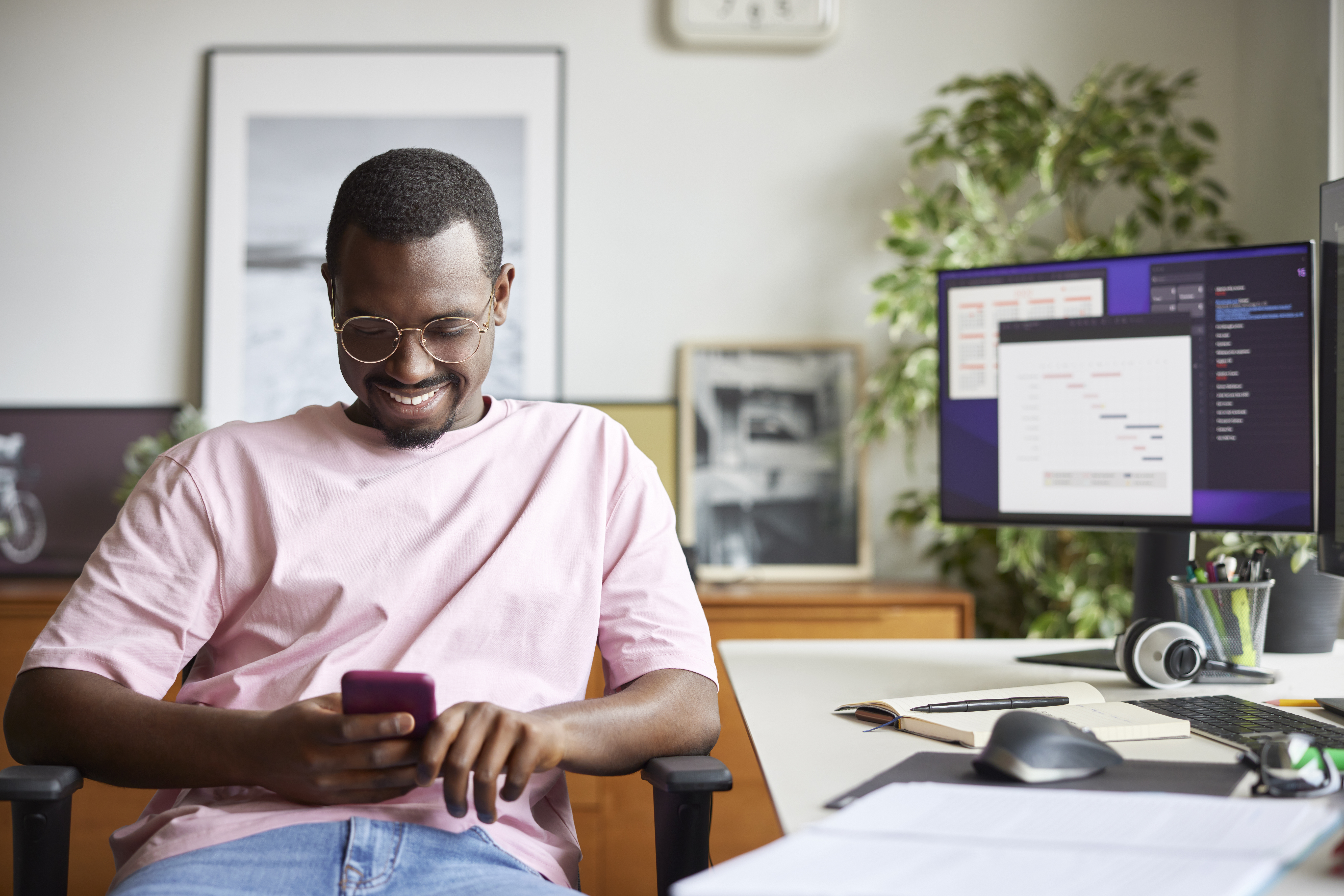 Smiling businessman using smart phone while sitting by computer desk. Male professional is surfing internet at home office. He is in casuals at apartment.