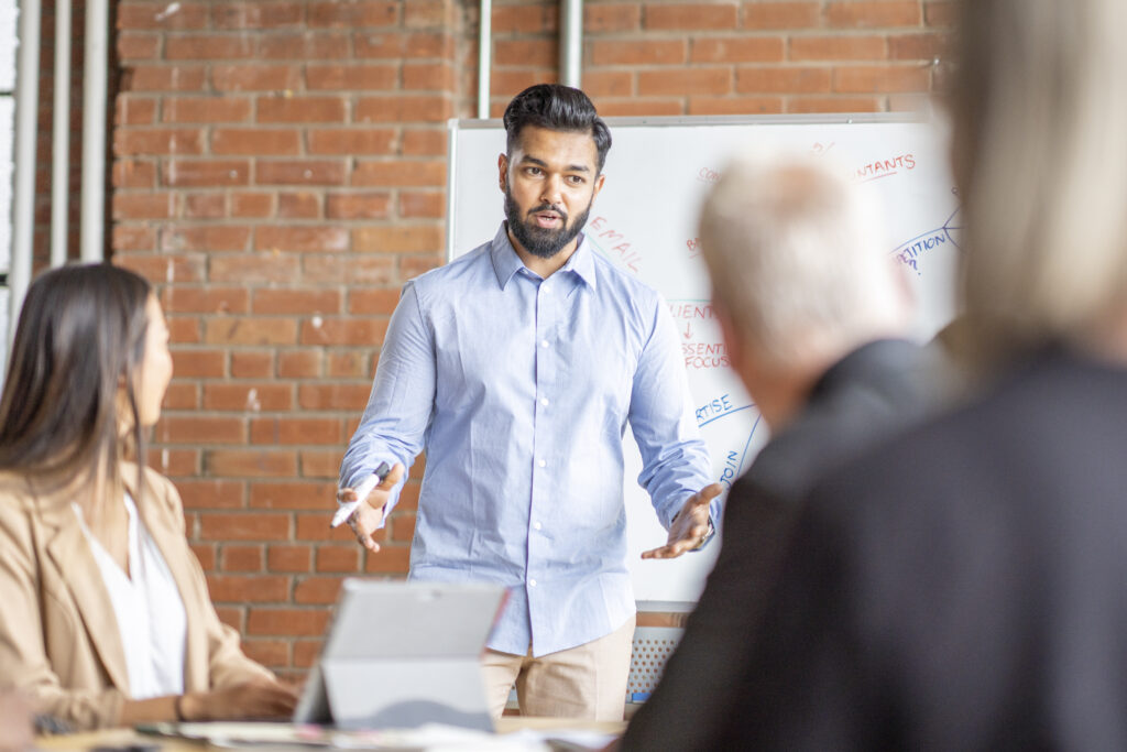 A young gentleman of Middle Eastern decent, stands at the head of a meeting table as he leads his team in a meeting. He is dressed professionally on a collared shirt and has his hands out to help him articulate his points about how HR drives revenue. His colleagues can be seen sitting at the table in front of him listening attentively.