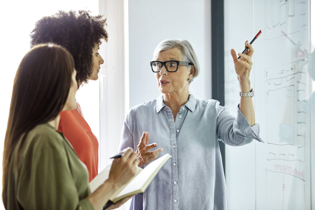 Senior female entrepreneur explaining the four-day workweek on glass wall to businesswomen. Colleagues are discussing in board room. They are working in office.